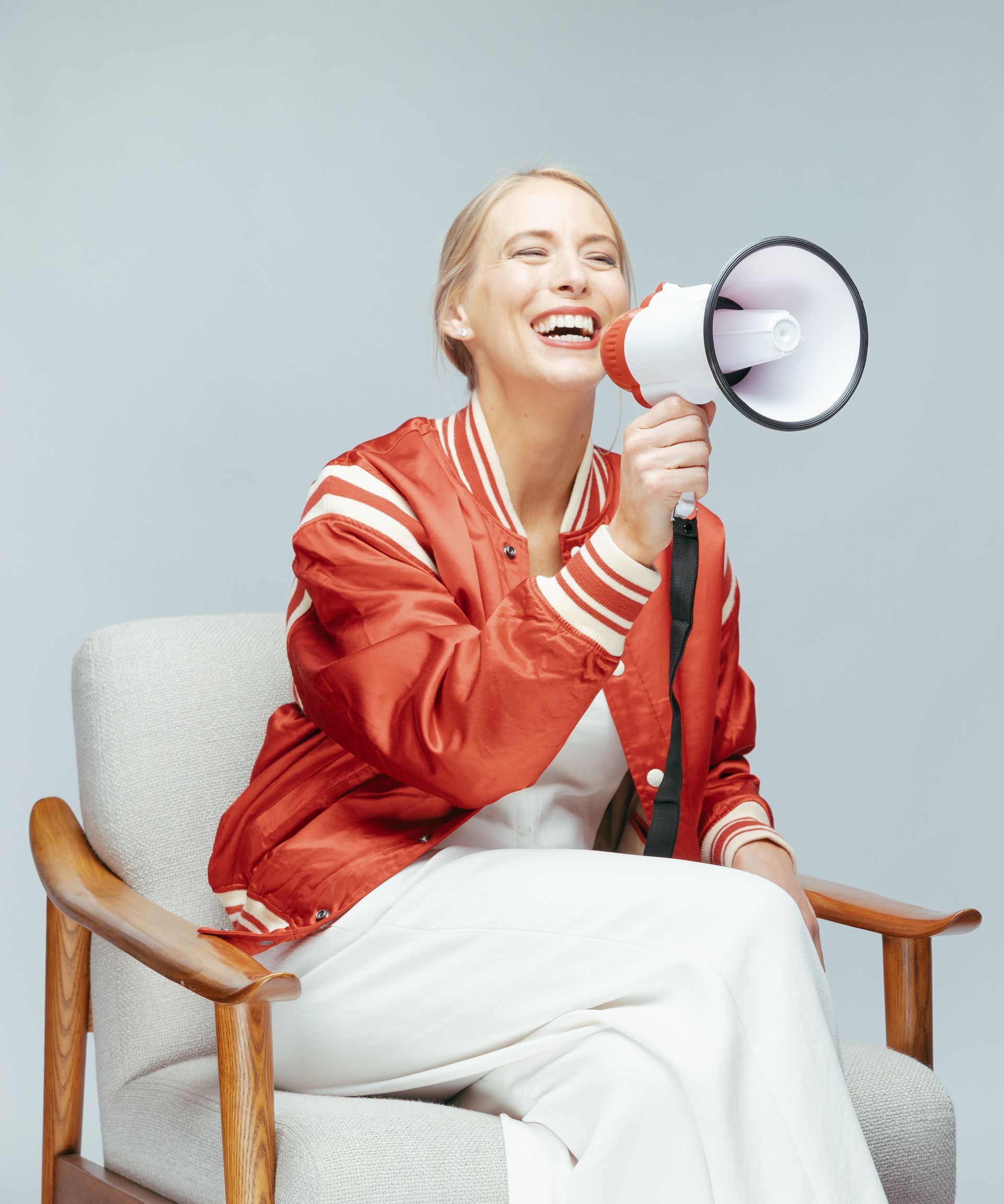Woman in red jacket, white outfit, sitting in chair, holding megaphone, laughing.
