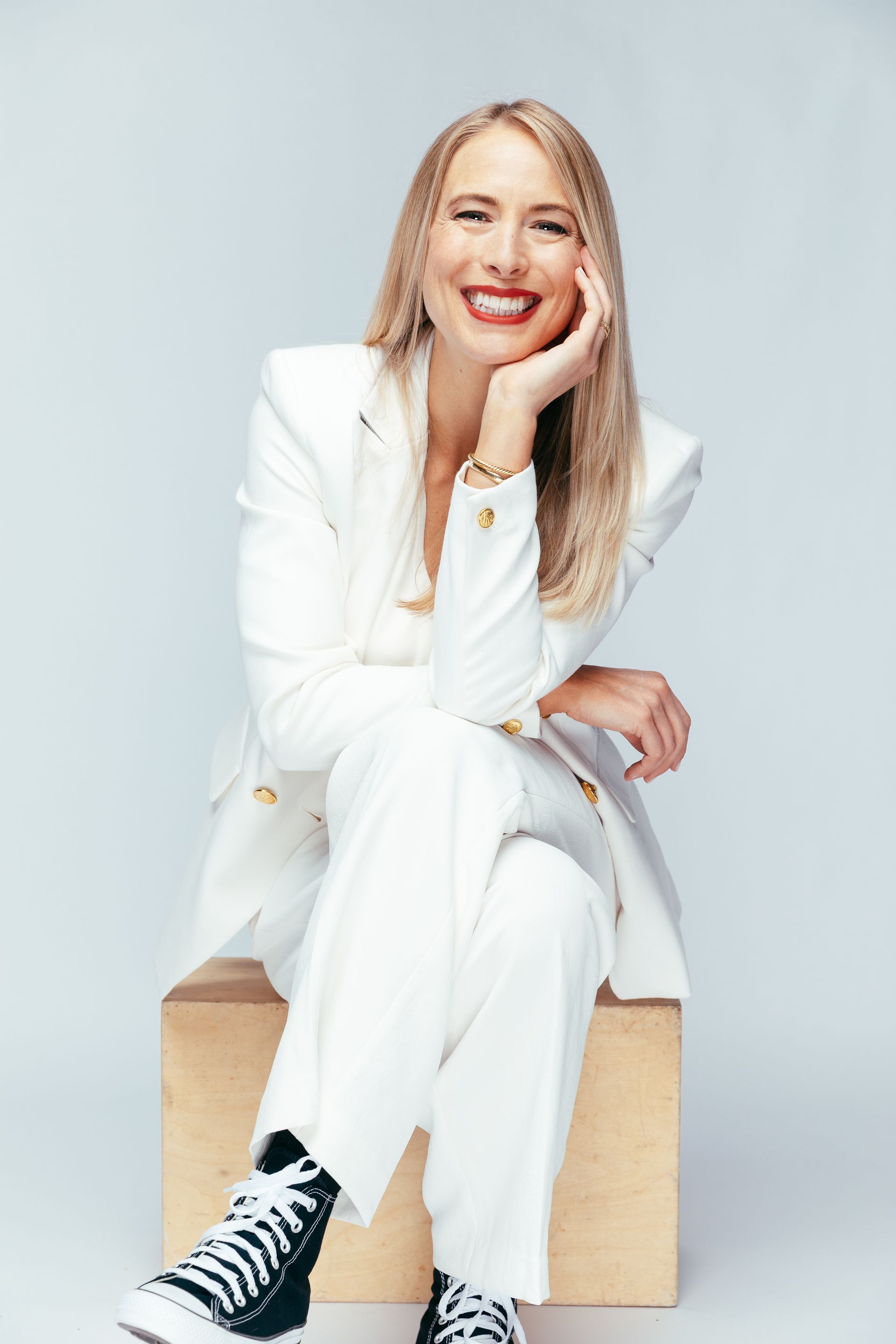 Woman in white suit and black high-tops, smiling, resting chin on hand, sitting on a wooden box.