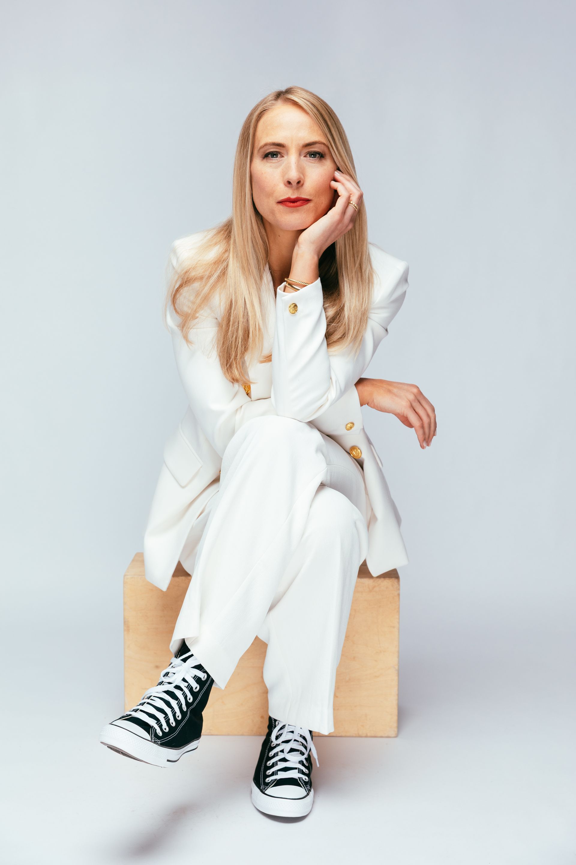 Woman in white suit and sneakers, seated on a box, hand to her chin, looking at the camera.