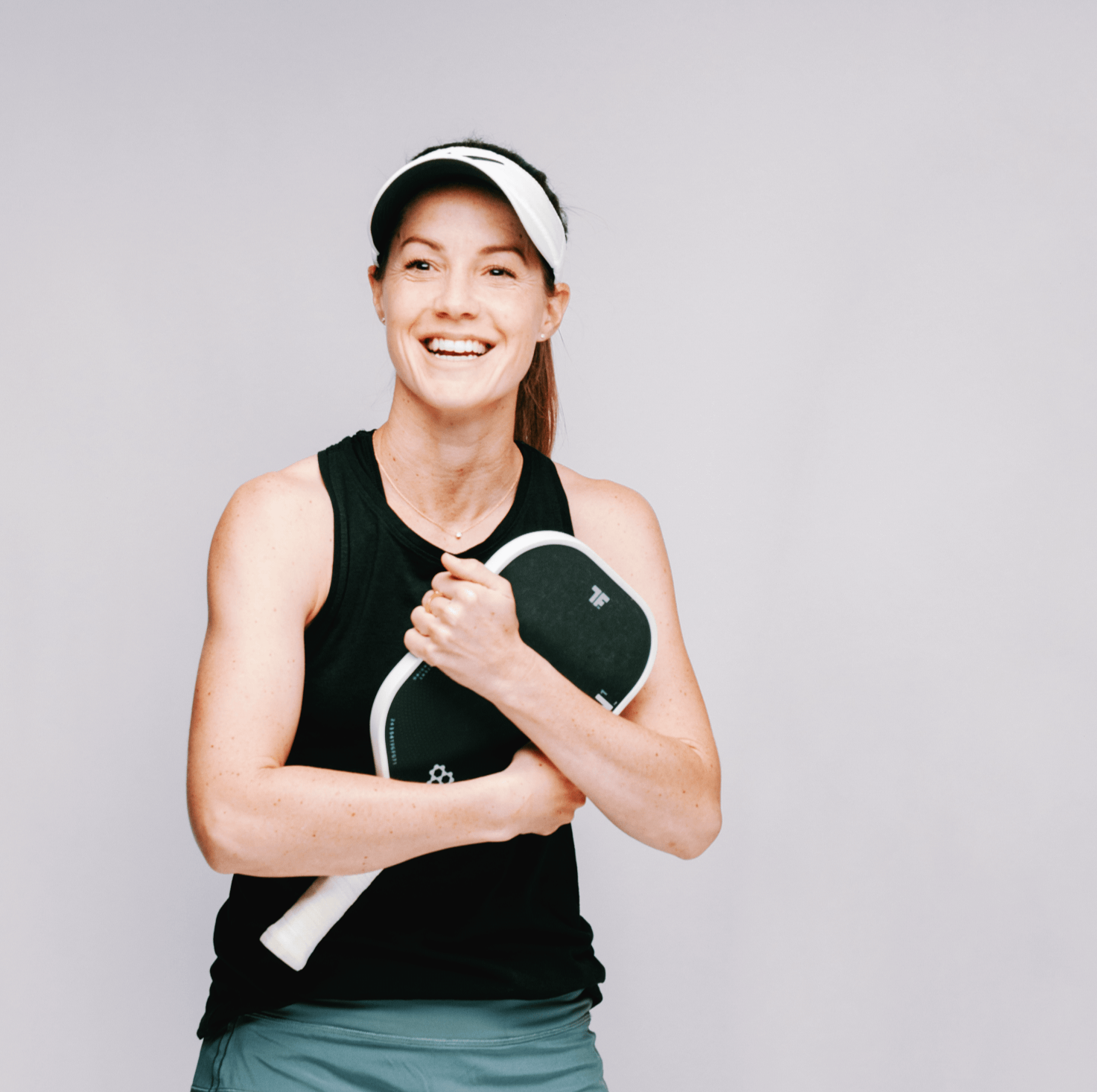 Woman in visor, holding a pickleball paddle, smiling against a neutral background.