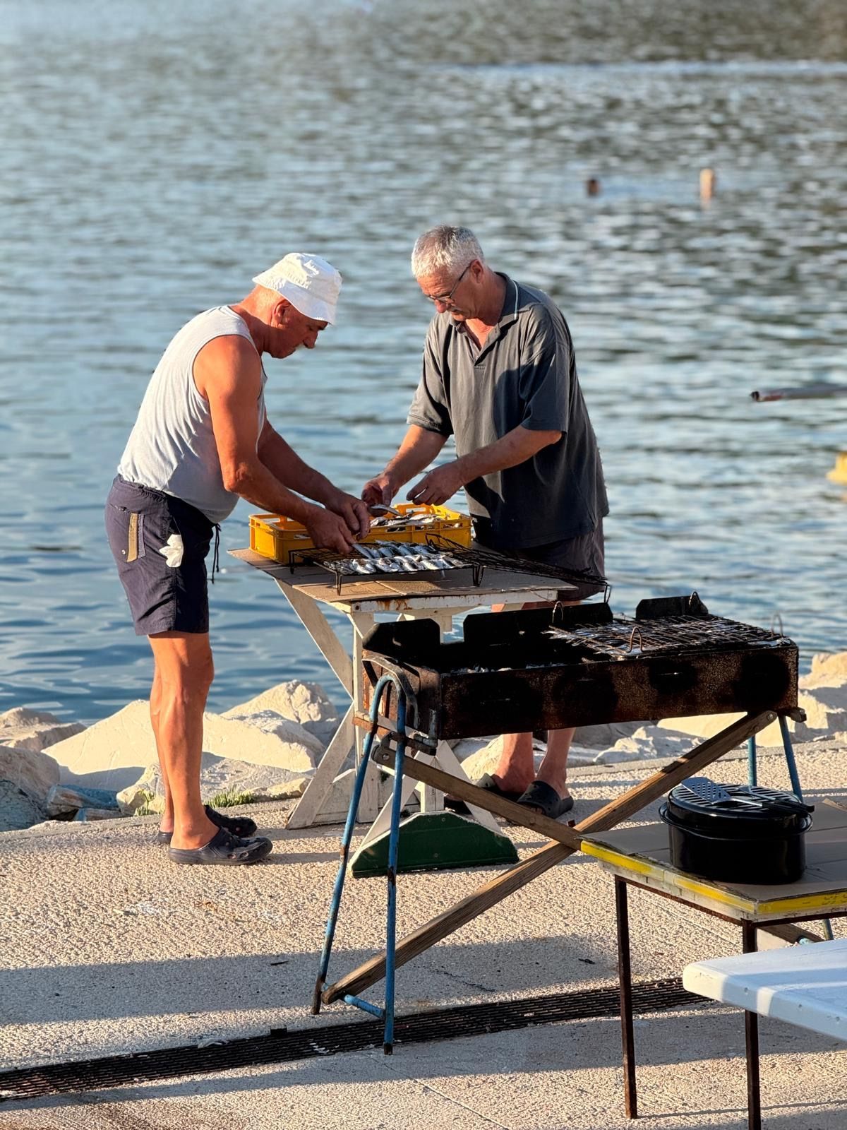 Two men grilling food by the water; one wears a white hat and tank top, the other a grey shirt.