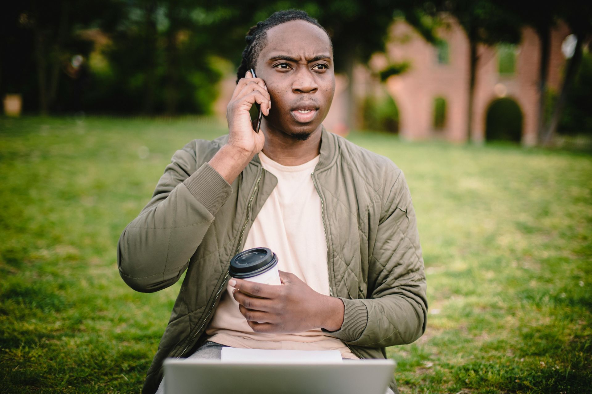 Man with dreadlocks on a phone call, looking concerned, holding coffee and using a laptop in a park.