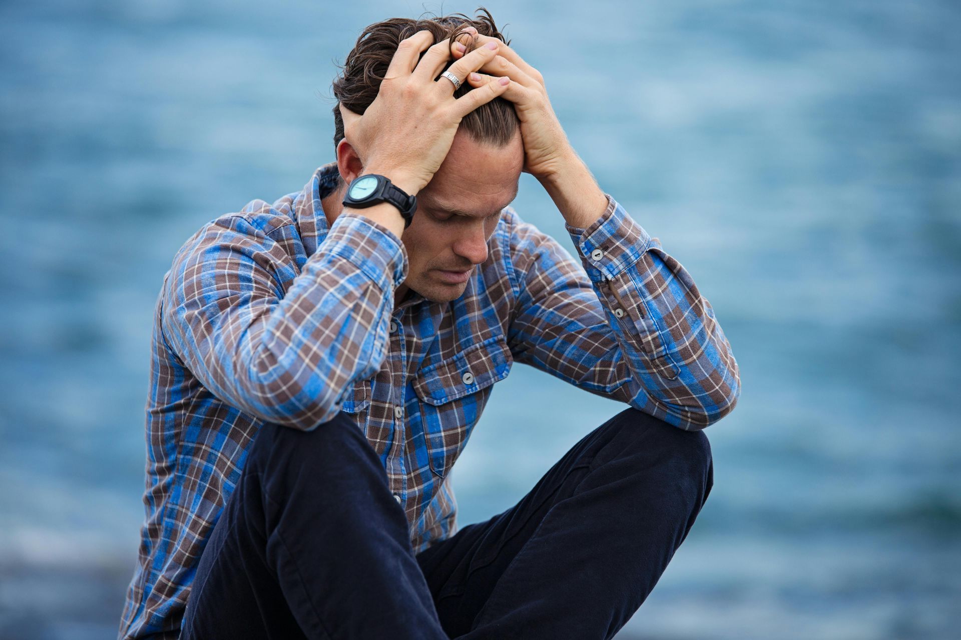 Man sitting with head in hands, looking down, showing distress. Outdoors, blue background.