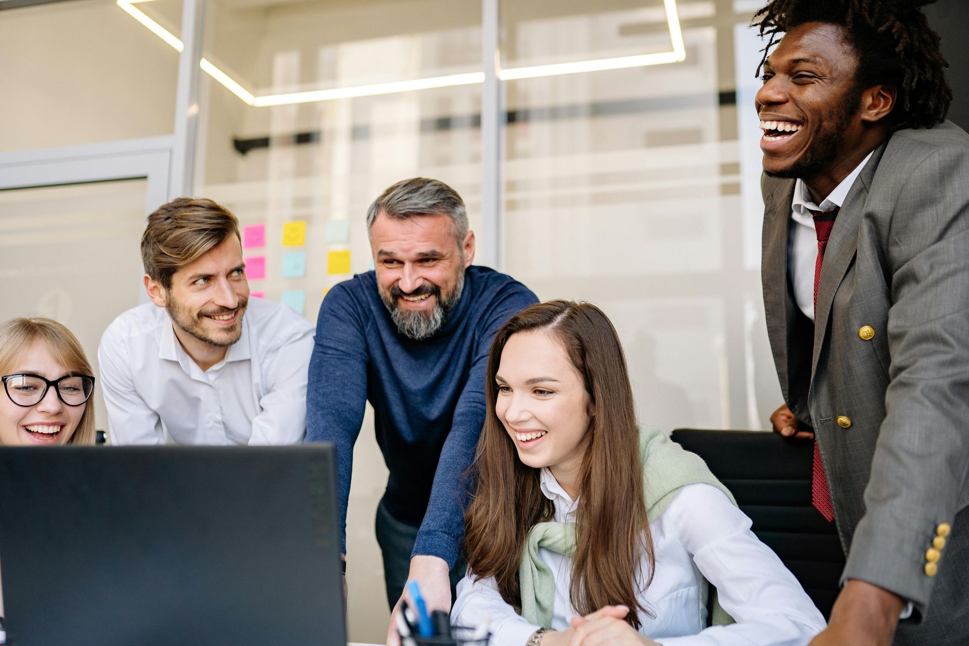 Group of diverse colleagues smiling and laughing while looking at a laptop in an office setting.