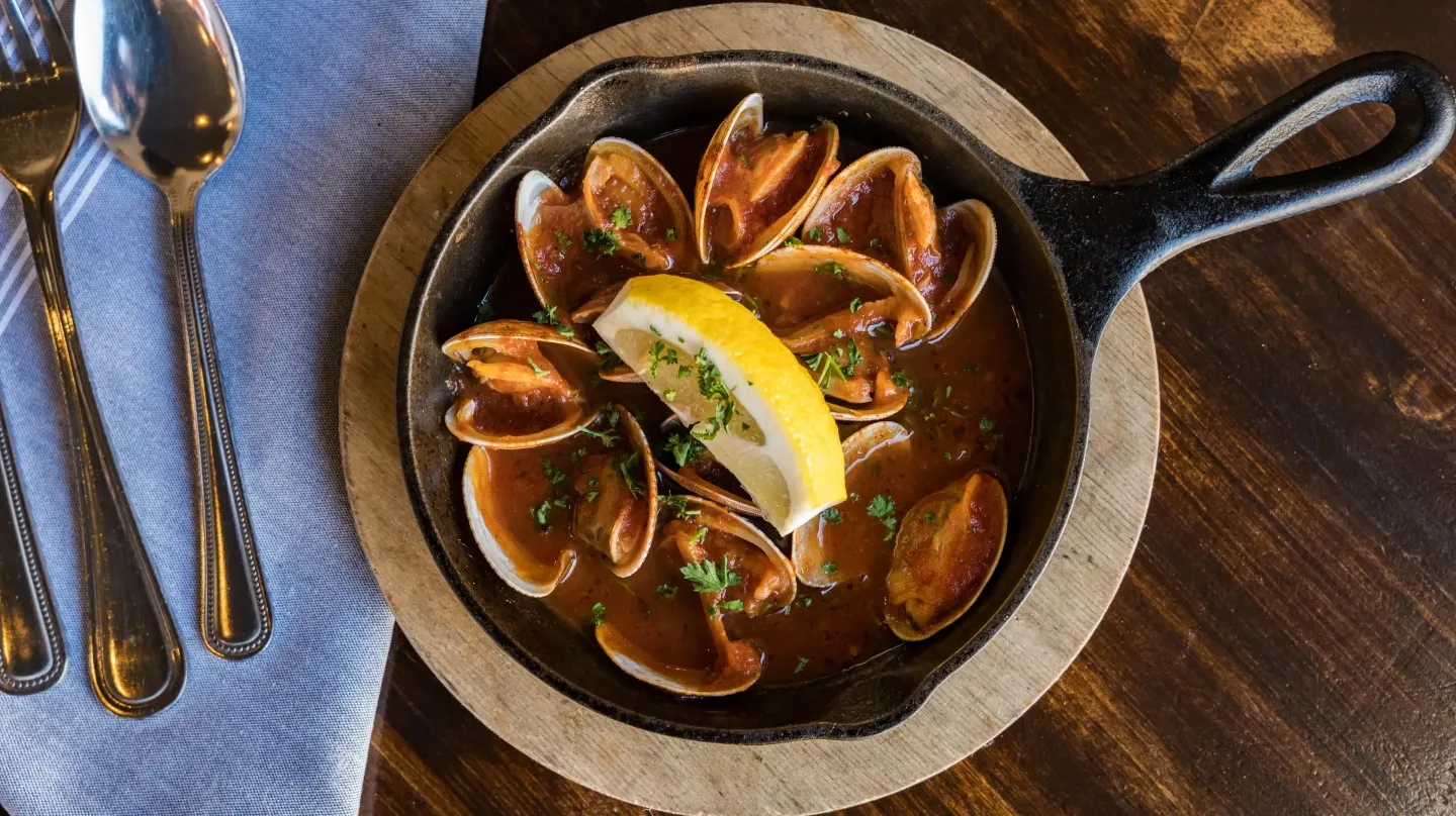 A skillet filled with clams and a slice of lemon on a wooden table.