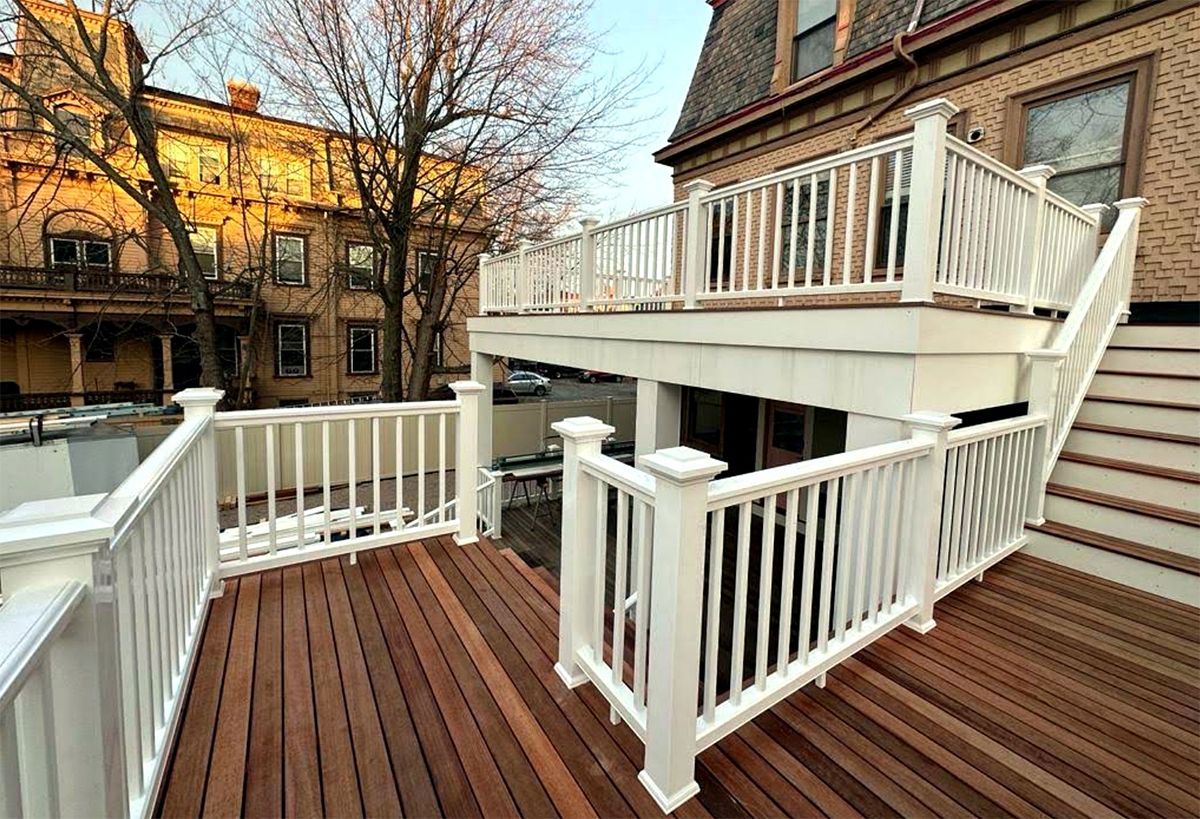 Wooden deck with white railings beside a brick townhouse, overlooking a courtyard with autumn trees
