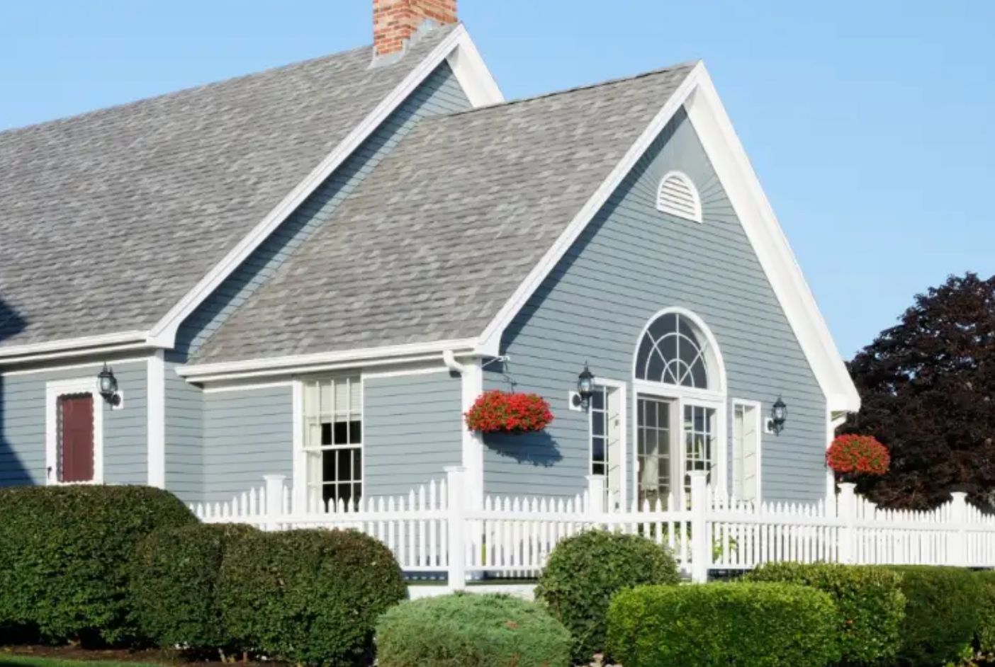 Gray house with white trim, arched window, and red flower baskets behind a white fence