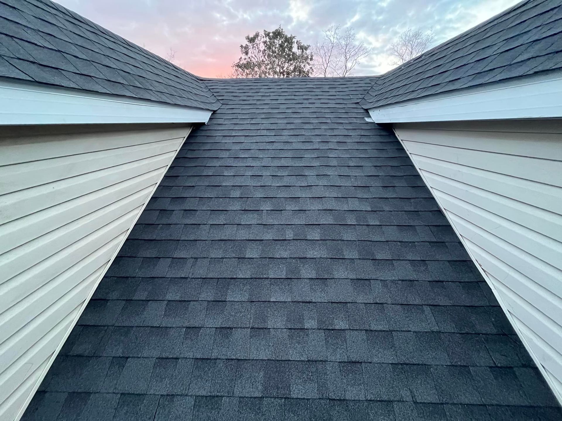 Steep dark gray shingled roof between two white house walls under a pink evening sky