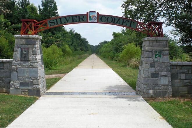 Stone entrance arch reading “Silver Comet” over a paved trail through green trees.
