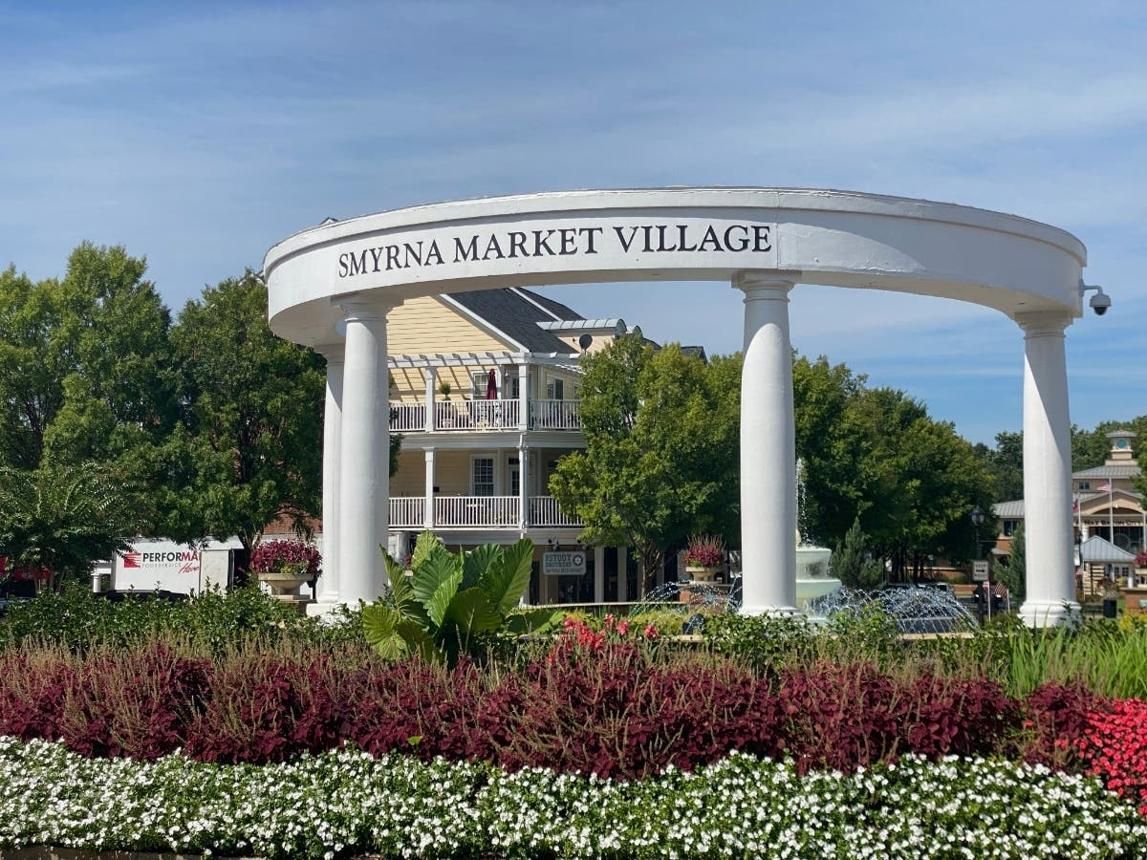 Elyria Market Village entrance sign beneath a white archway, with flowers and trees in front of shops.