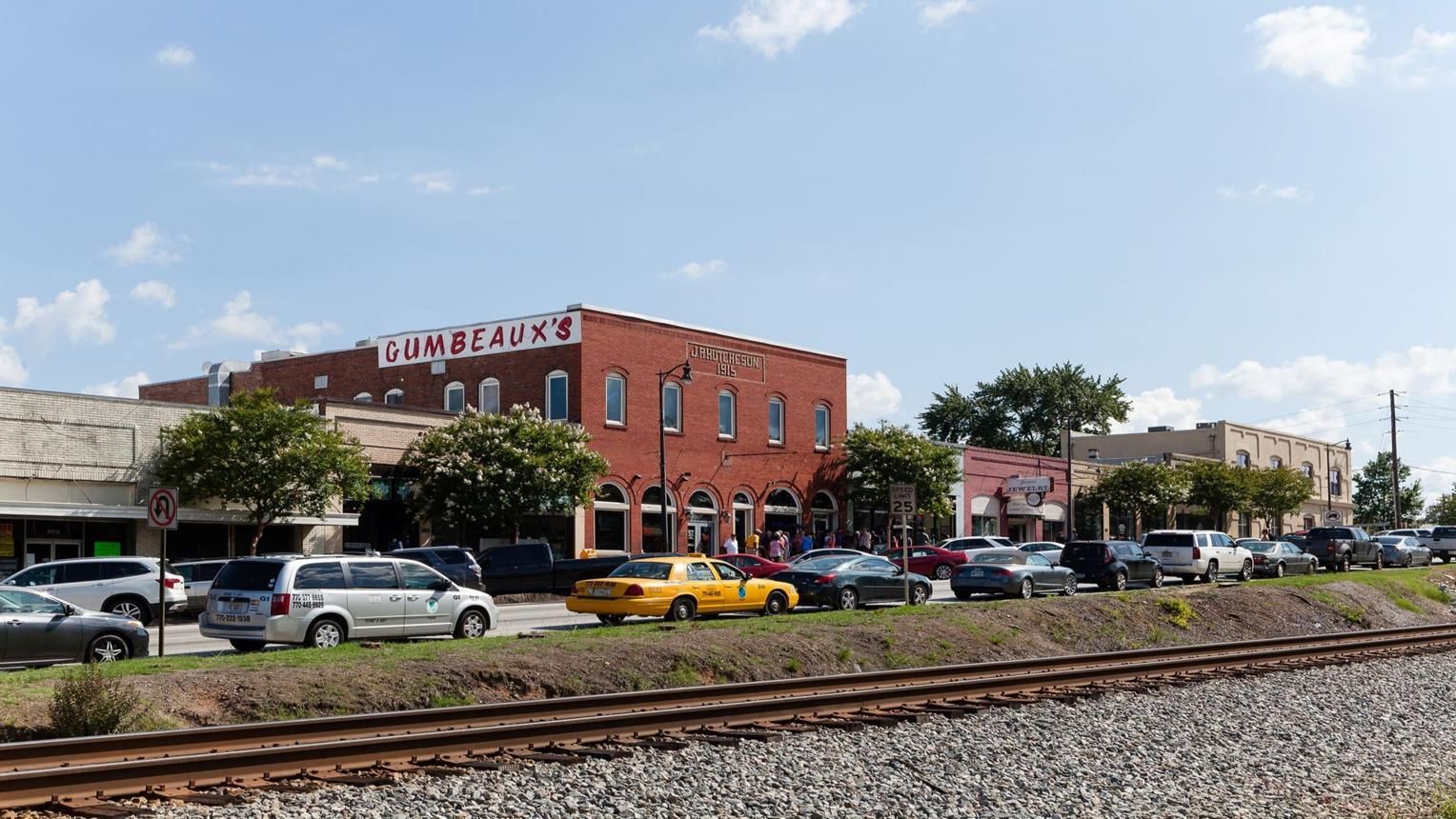 Street with railroad tracks, parked cars, and a red-brick building under a partly cloudy sky