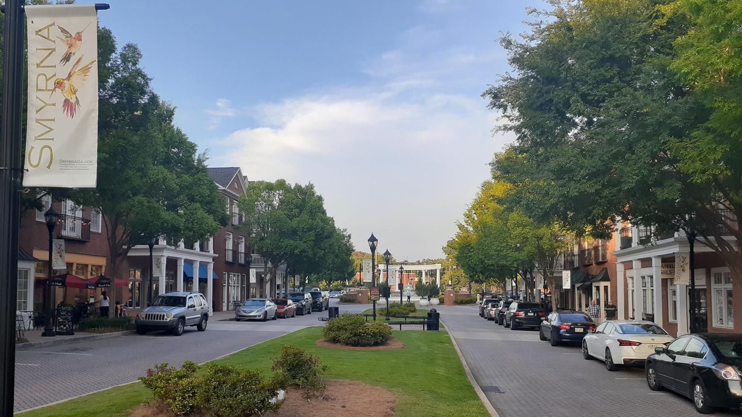 Tree-lined city street with parked cars, a grassy median, and low-rise buildings under a partly cloudy sky