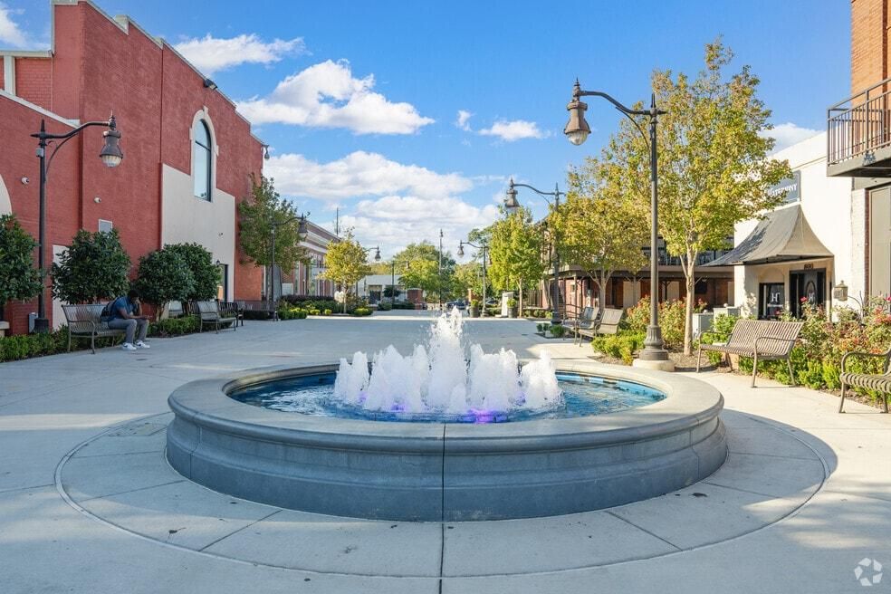 Circular fountain in a sunny plaza with red brick buildings, trees, and blue sky