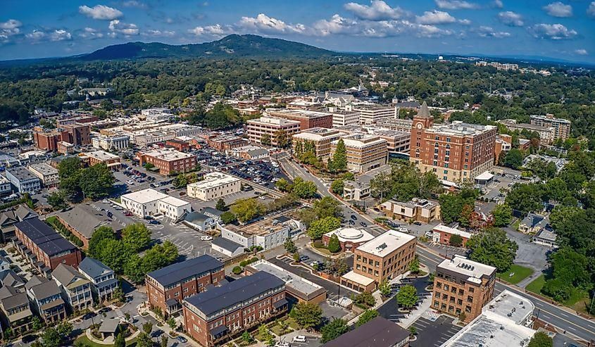 Aerial view of a small city with downtown buildings, tree-lined streets, and mountains under a blue sky.
