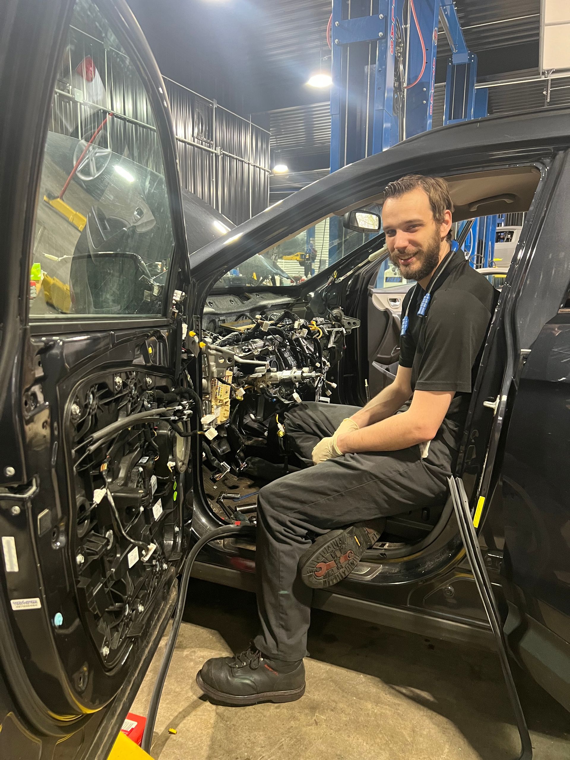 Man sitting in a black car frame on an assembly line, smiling in a factory setting | Pro Import Automotive Service and Repair