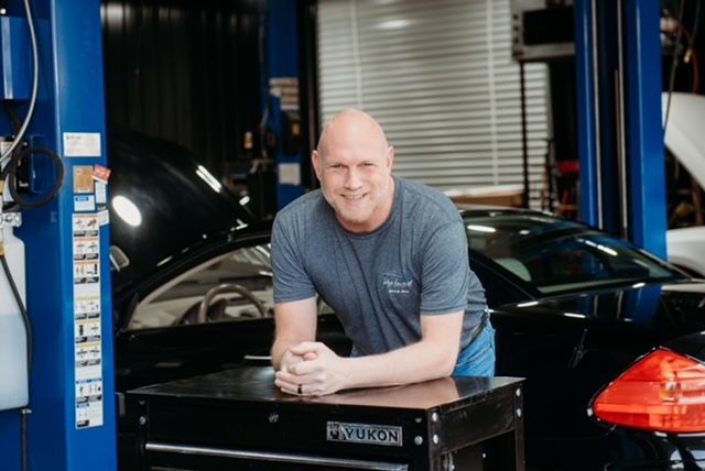 Mechanic leaning on a tool cart in an auto repair shop beside a black car | Pro Import Automotive Service and Repair