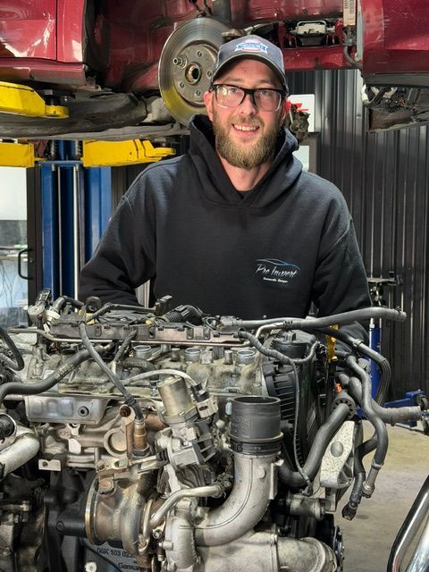 Mechanic smiling in a workshop, standing beside a large engine and car lift. | Pro Import Automotive Service and Repair