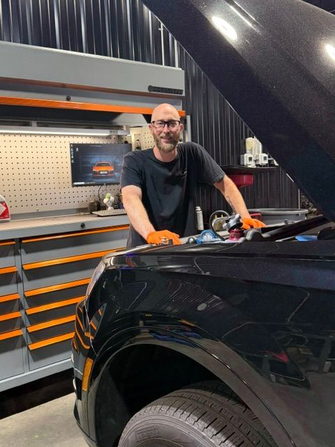 Mechanic smiling beside a black car in a workshop, with tools and orange storage | Pro Import Automotive Service and Repair