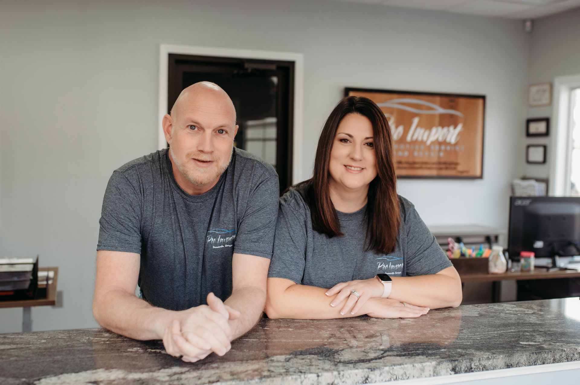 A man and woman in matching grey shirts leaning on a granite counter in an office. | Pro Import Automotive Service and Repair