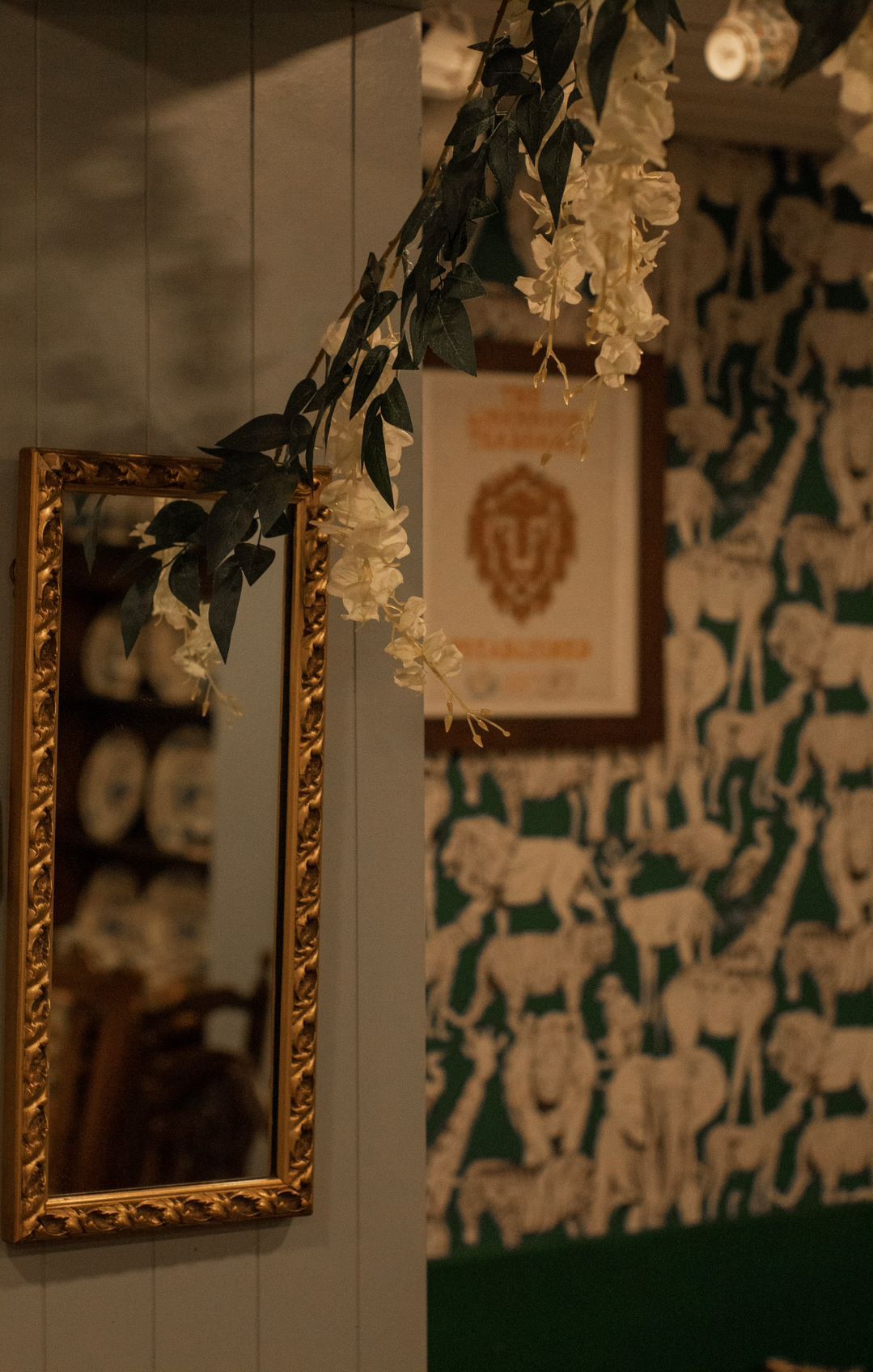 Gold framed mirror and printed artwork hanging on wall with animal-patterned wallpaper. White flower garland draped.
