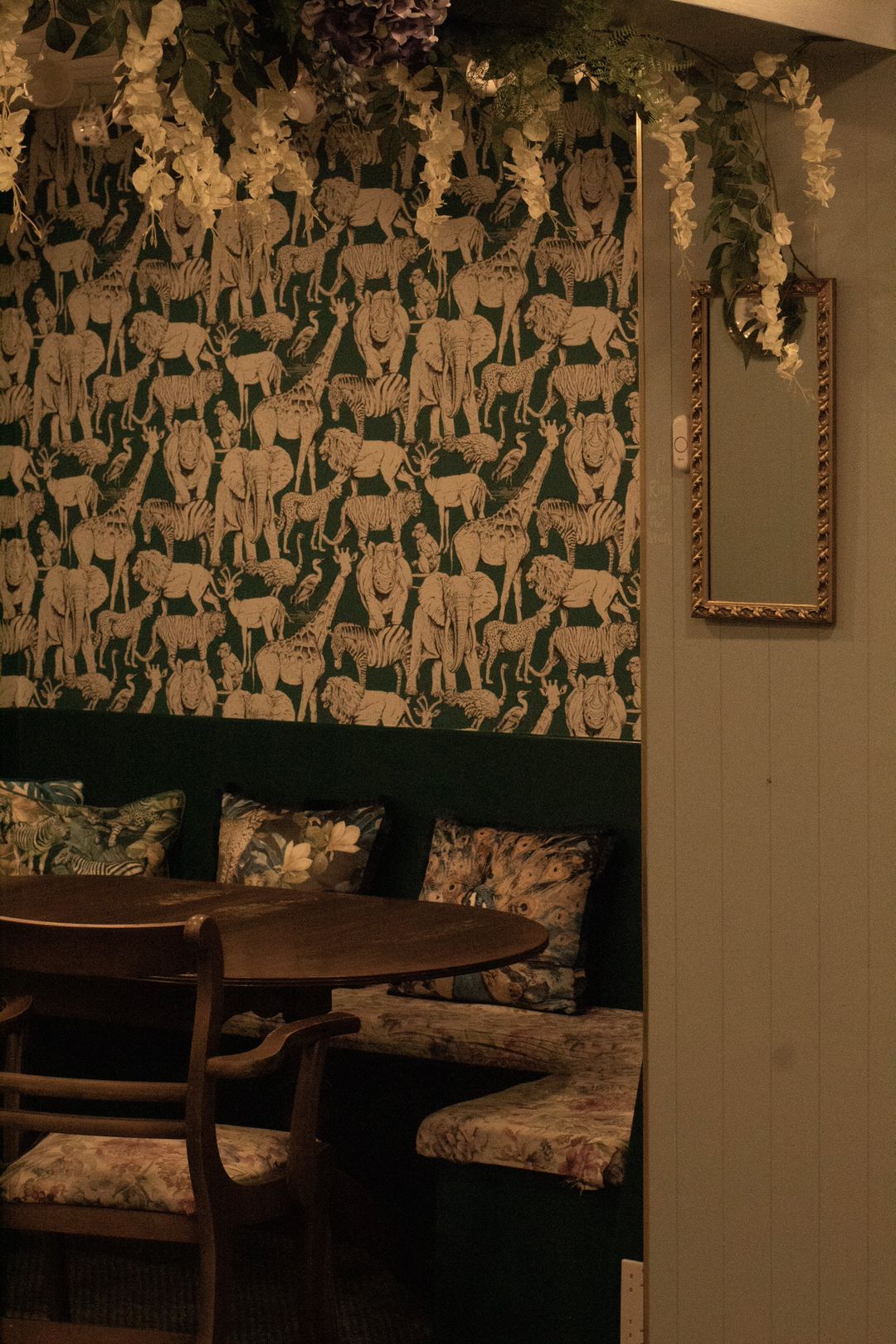 Cozy dining nook with green wallpaper and botanical decor. Round table, cushioned bench, and mirror.