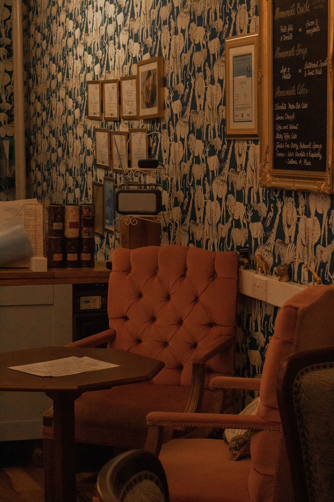 Cozy café corner with orange tufted armchairs, a small table, patterned wallpaper, and framed menus.