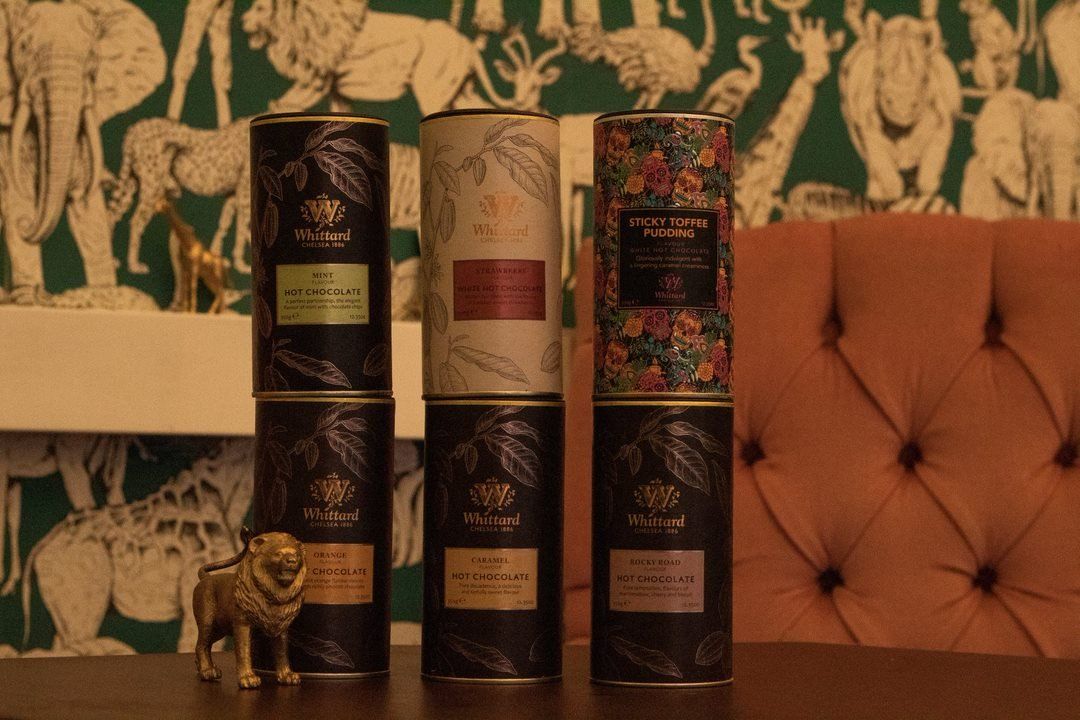 Six decorative tea canisters stacked on a table, with a gold lion statue, against a patterned wallpaper.