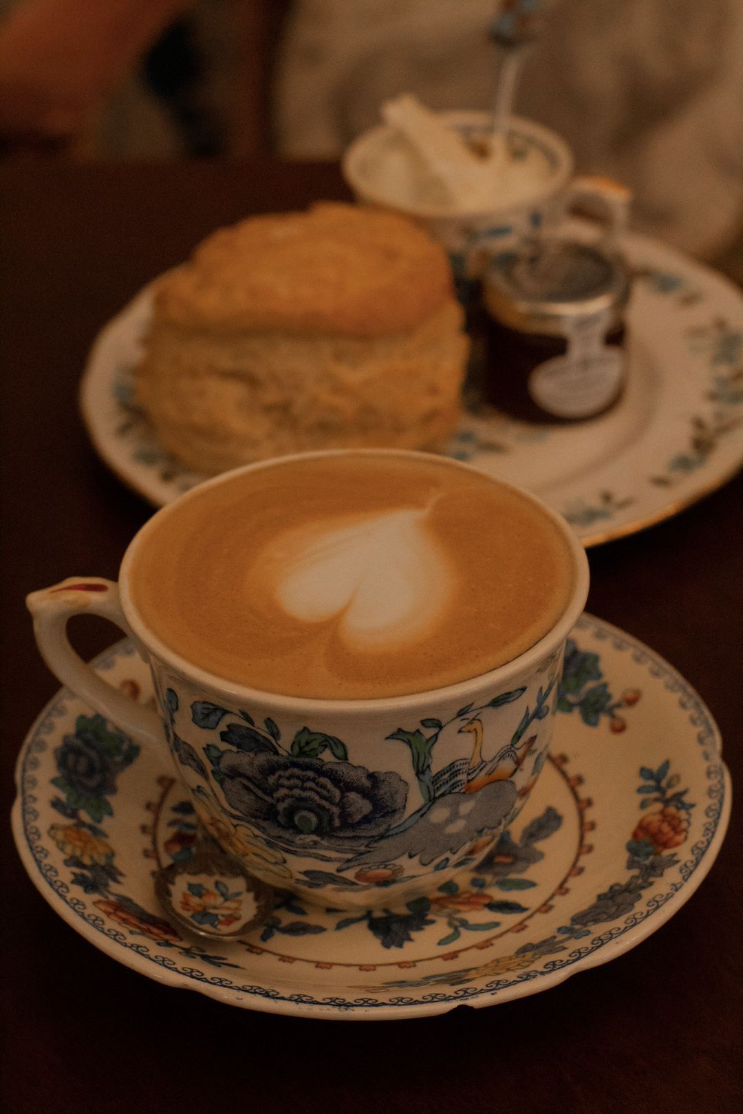 Cup of latte with heart design, scone, cream, and jam on a floral plate.