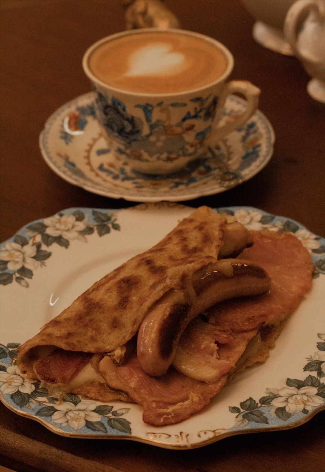 Breakfast plate with bacon, sausage, and a folded pancake; teacup with latte.