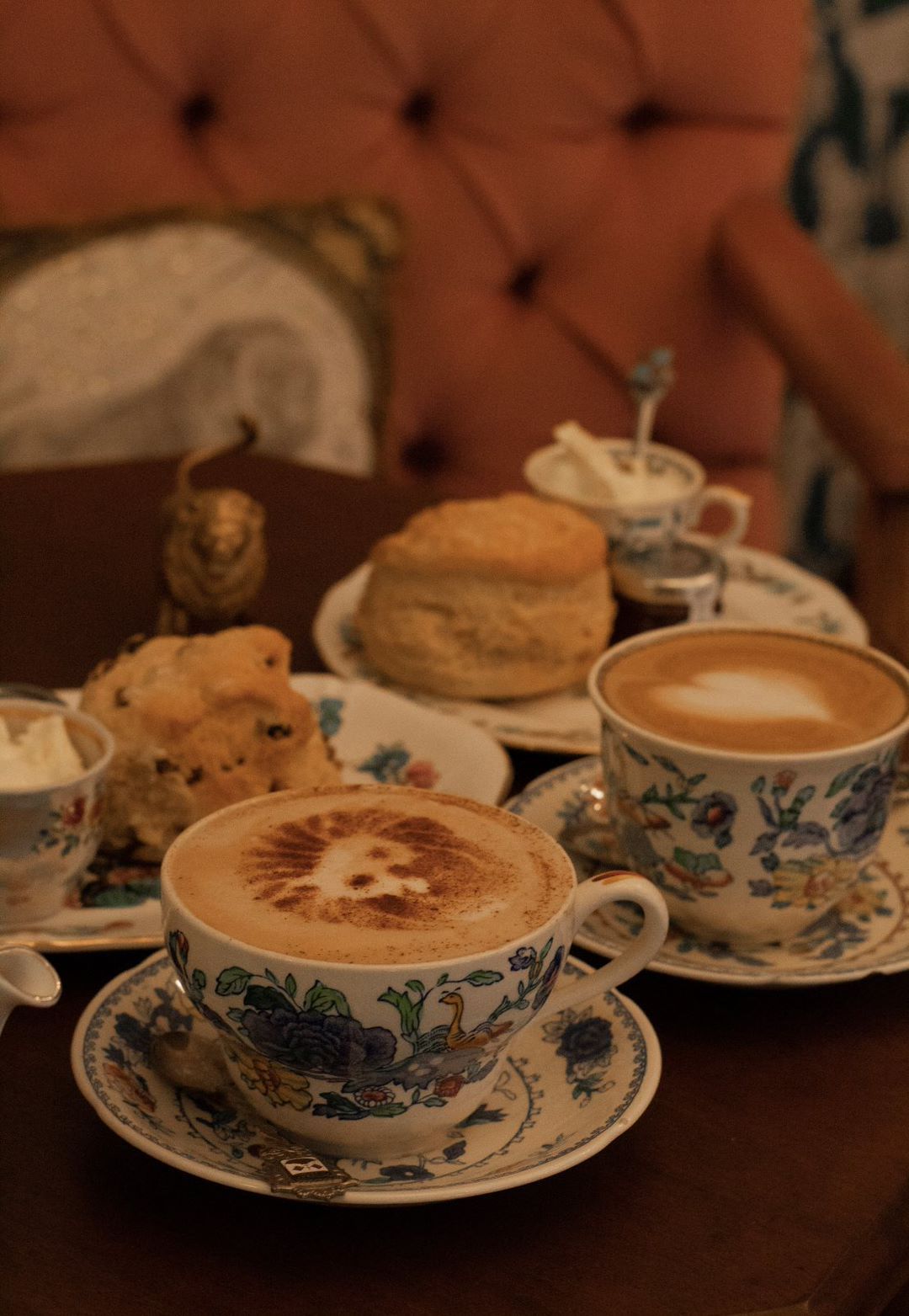 Two floral teacups of latte, scones, and cream on a wooden table. Pink tufted chair in background.
