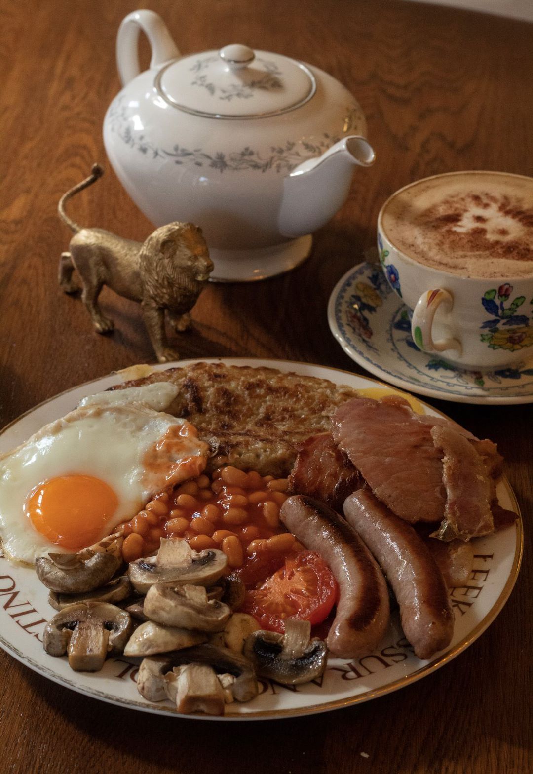 Full English breakfast on a plate with coffee, teapot, and a lion figurine on a wood table.