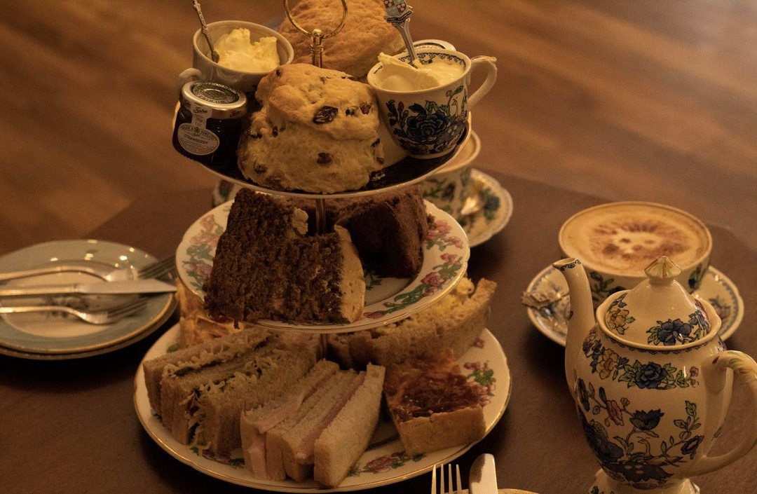 Three-tiered tea stand with scones, sandwiches, and cake, plus teacup, teapot, and coffee cup on table.