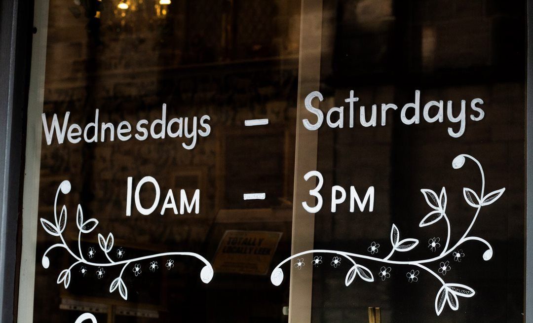 White lettering on a glass door: business hours are Wednesdays and Saturdays, 10 AM to 3 PM. Decorative floral accents.