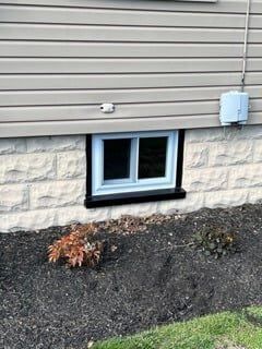 House foundation with a basement window, beige siding, stone veneer, gravel bed, and an electrical box.
