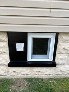 Basement window with white frame, black surround, and small vent under beige siding.