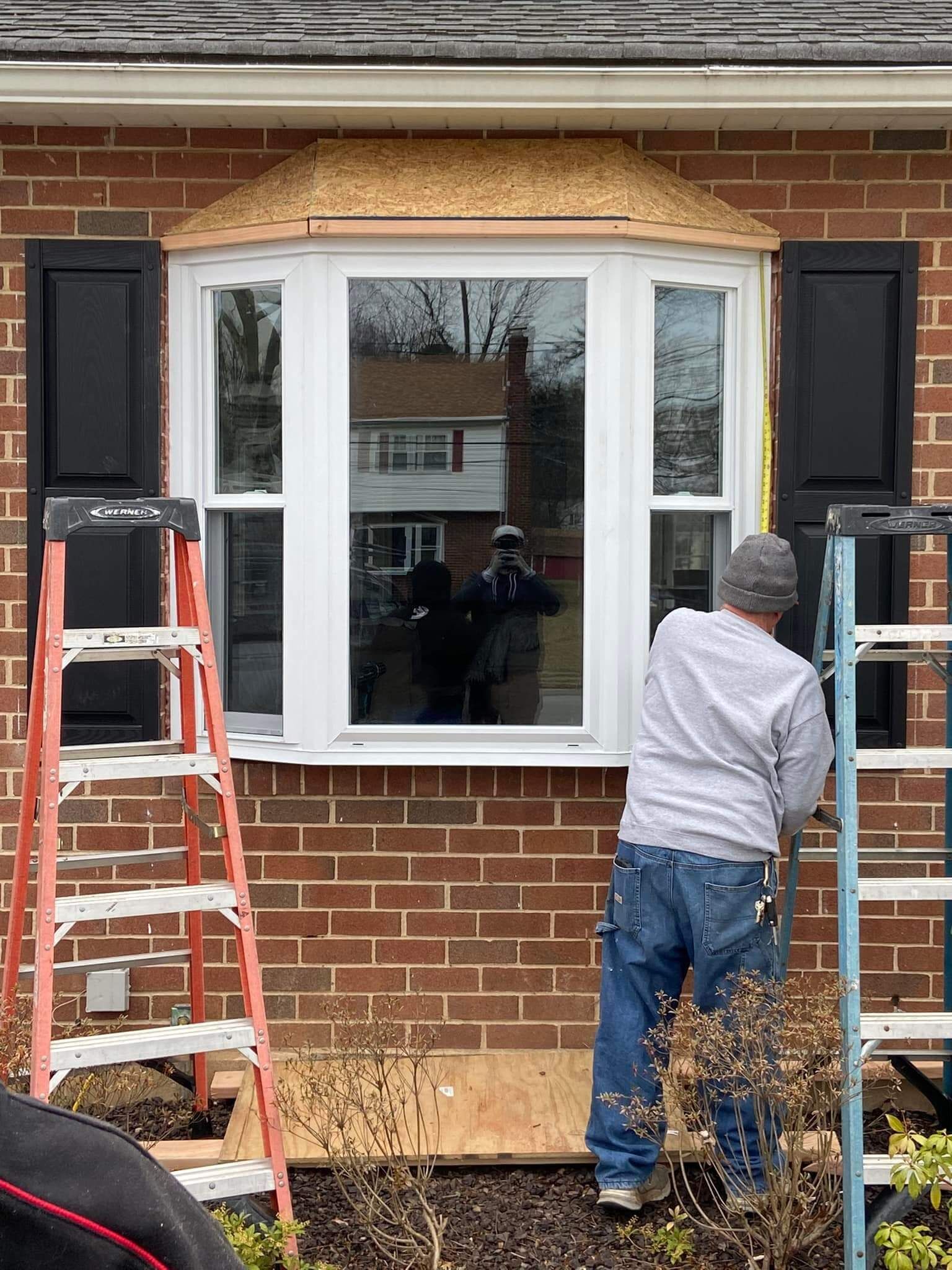 Worker installing trim around a white bay window on a brick house, with a ladder beside him.