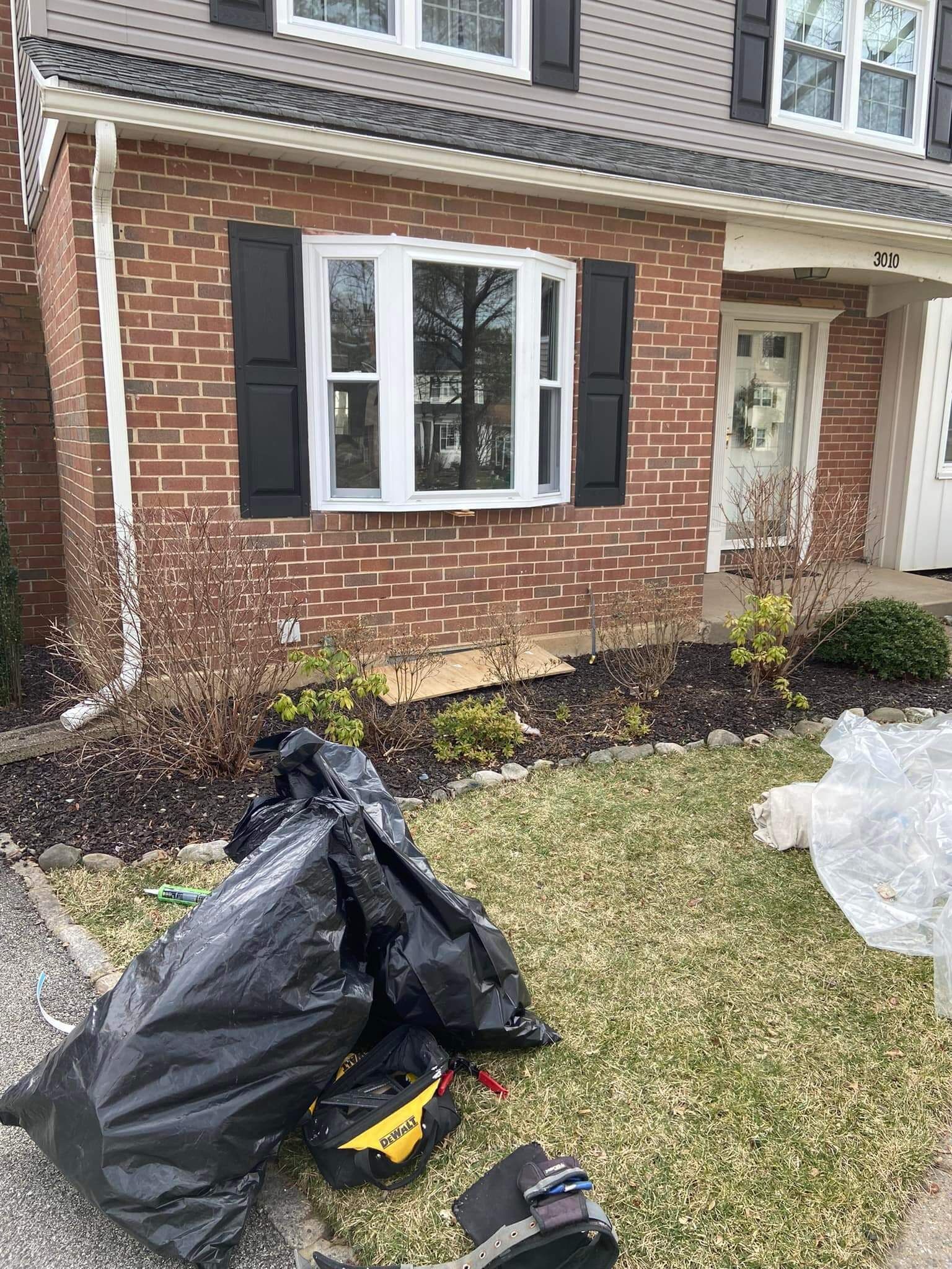 Brick townhouse entry with black shuttered window, front lawn debris bag, and a yellow tool on the grass