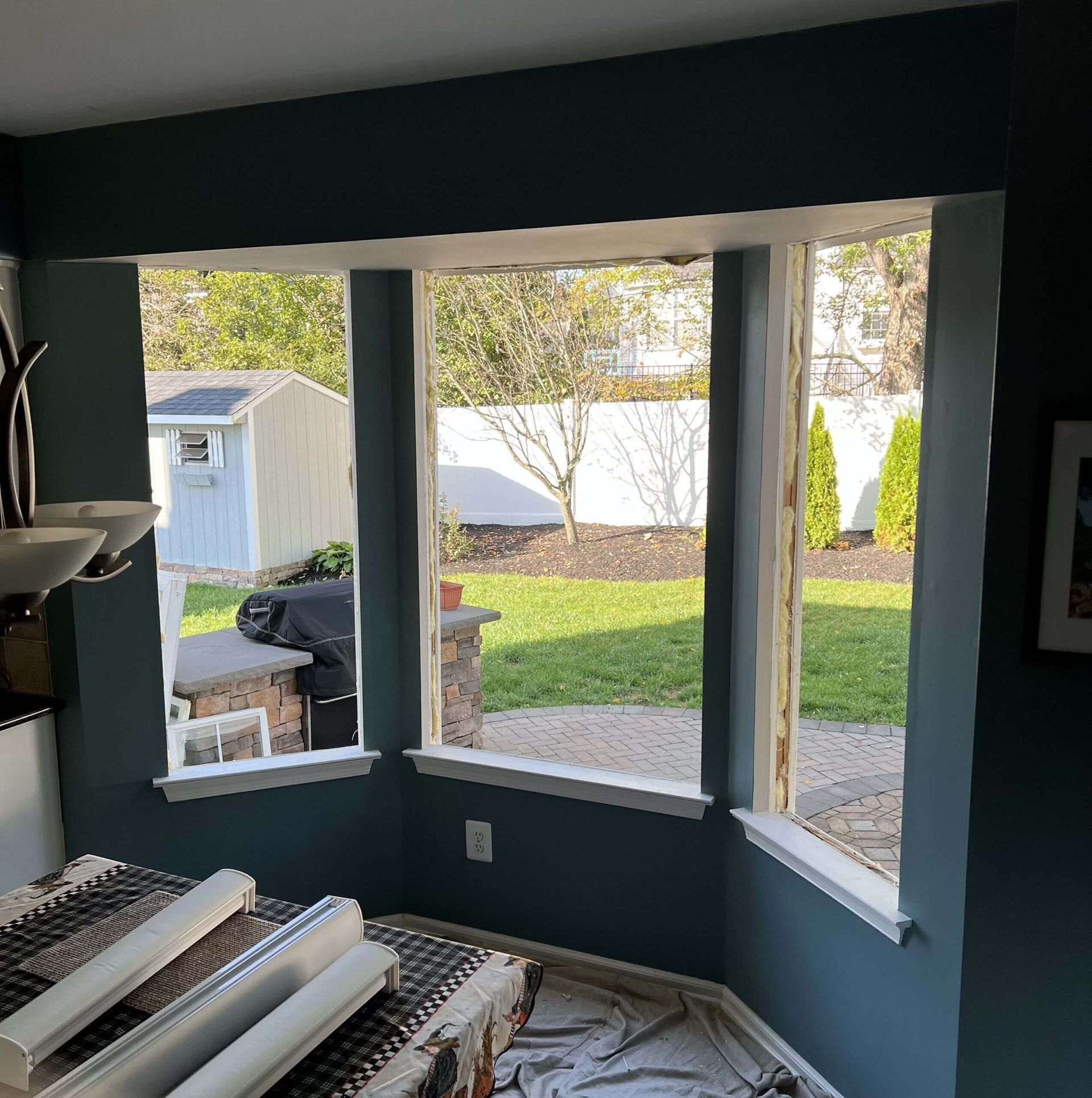 Living room bay window view of a yard with a shed, grill, and tree in daylight