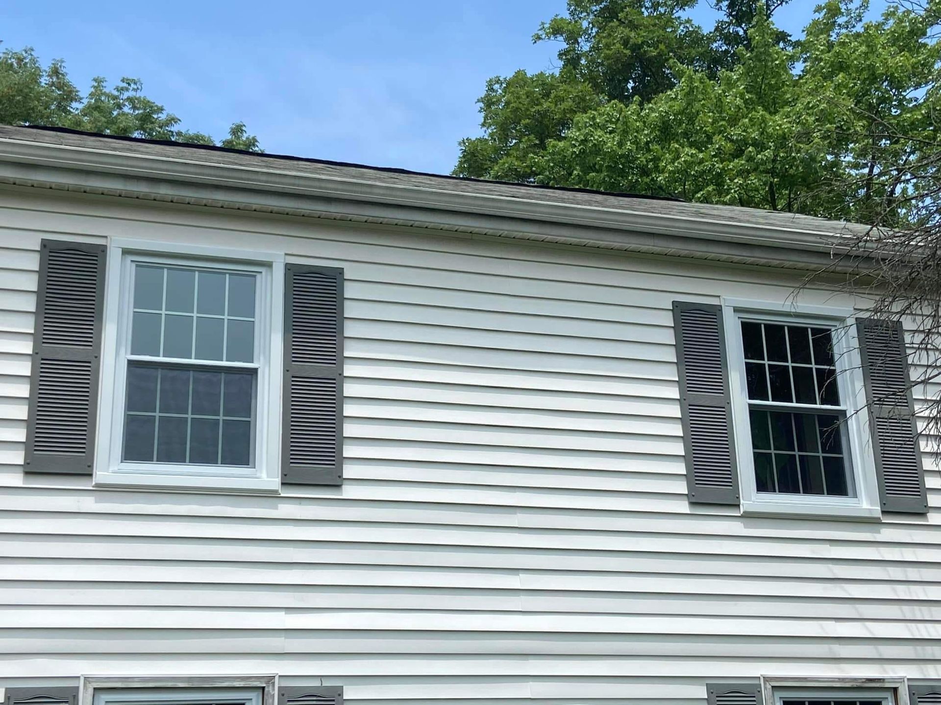 White house siding with two windows and gray shutters under a blue sky and green trees