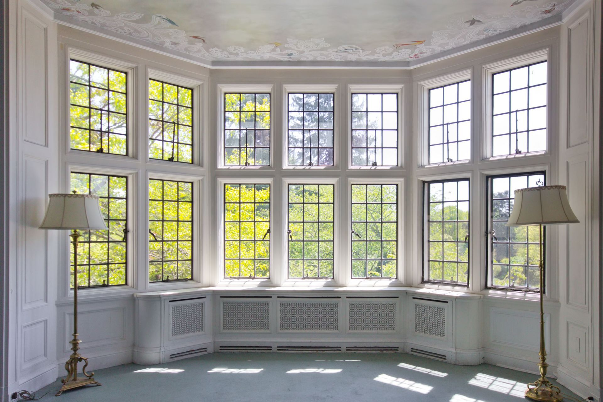 Curved bay window room with tall white-paned windows, floral ceiling, and two floor lamps
