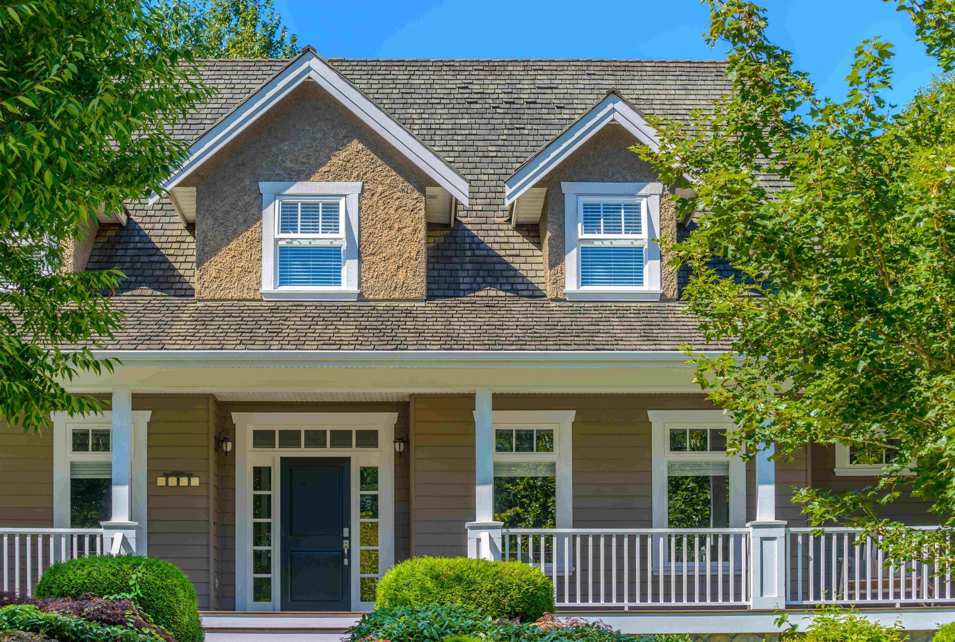 Brick house with a front porch, white railings, dormer windows, and green trees framing the entrance