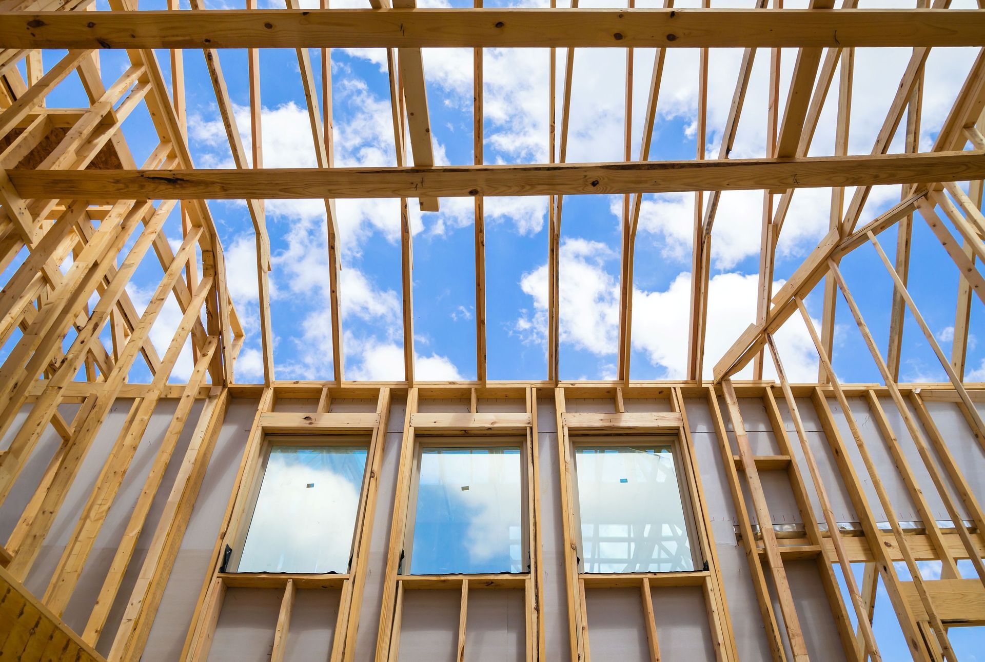 Wood-framed house under construction, viewed upward through open roof trusses against a blue sky.