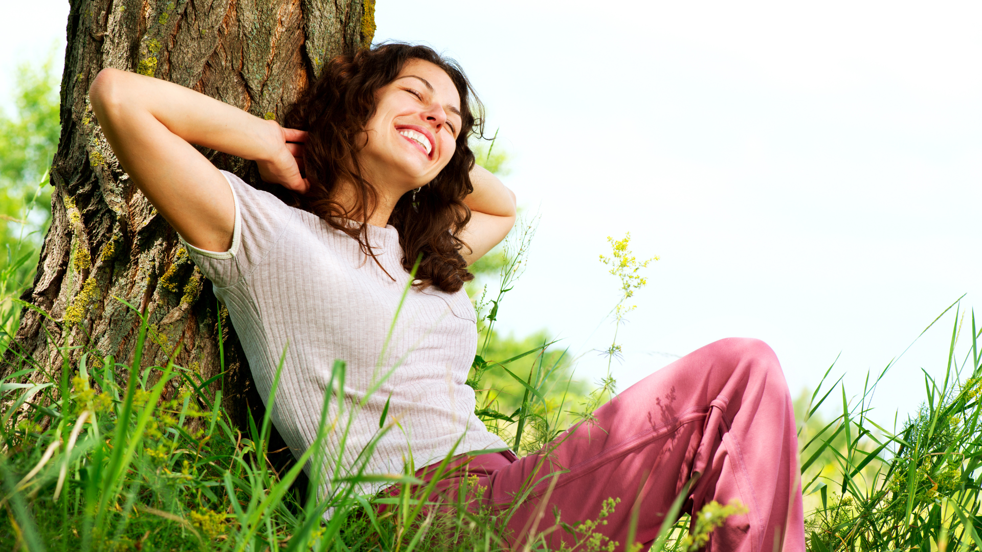 A woman is leaning against a tree in the grass and smiling.