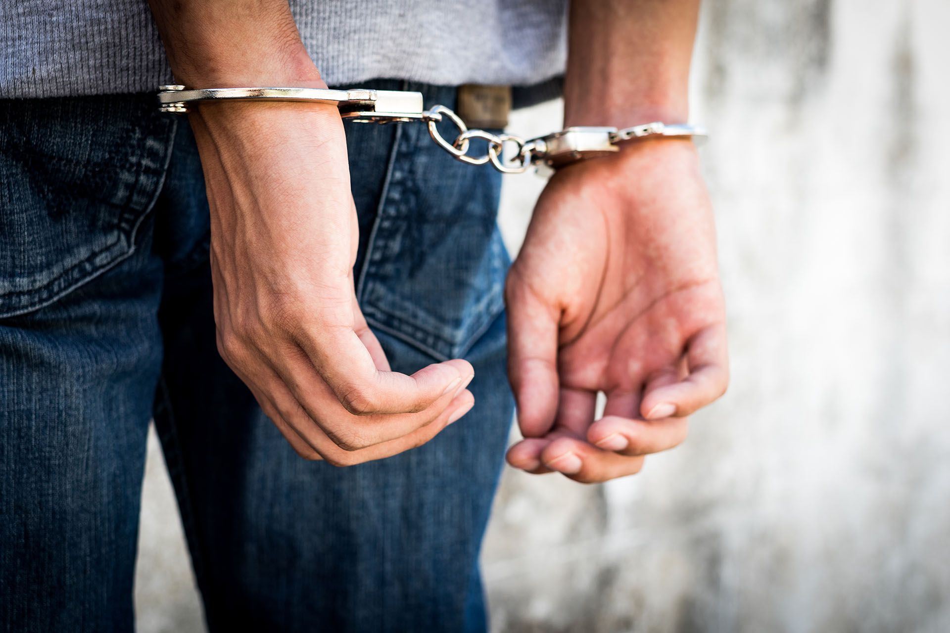 Person in handcuffs, wearing blue jeans and a gray sweater, against a textured wall.