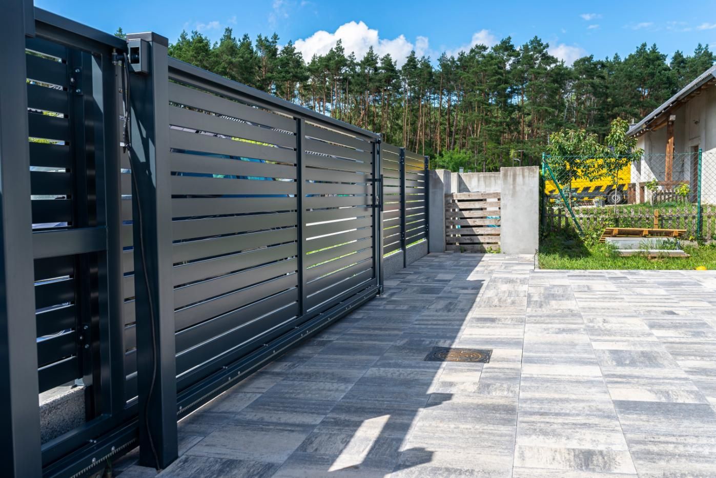 Black sliding driveway gate, gray brick pavers, house, trees, and blue sky.