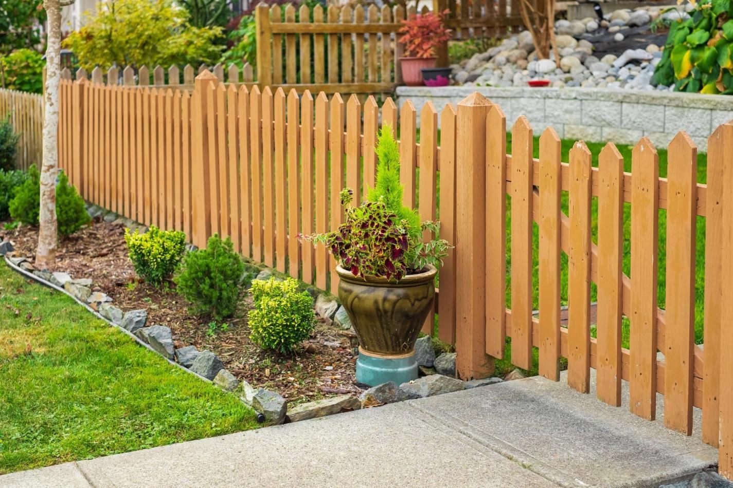 Wooden picket fence bordering a garden with green grass, shrubs, and a flower pot.