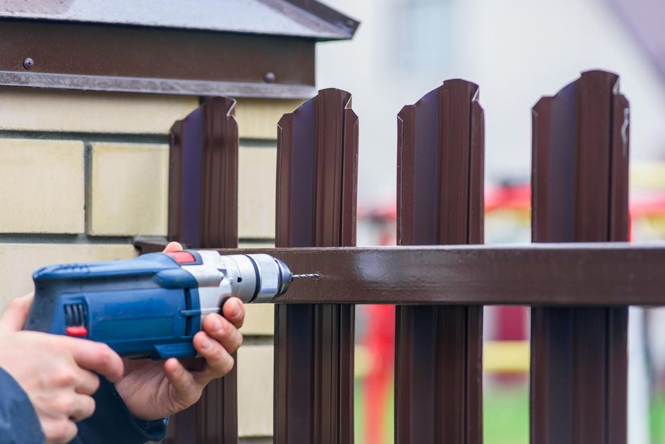 Person using a blue drill to attach a brown metal fence to a brown fence post outside.