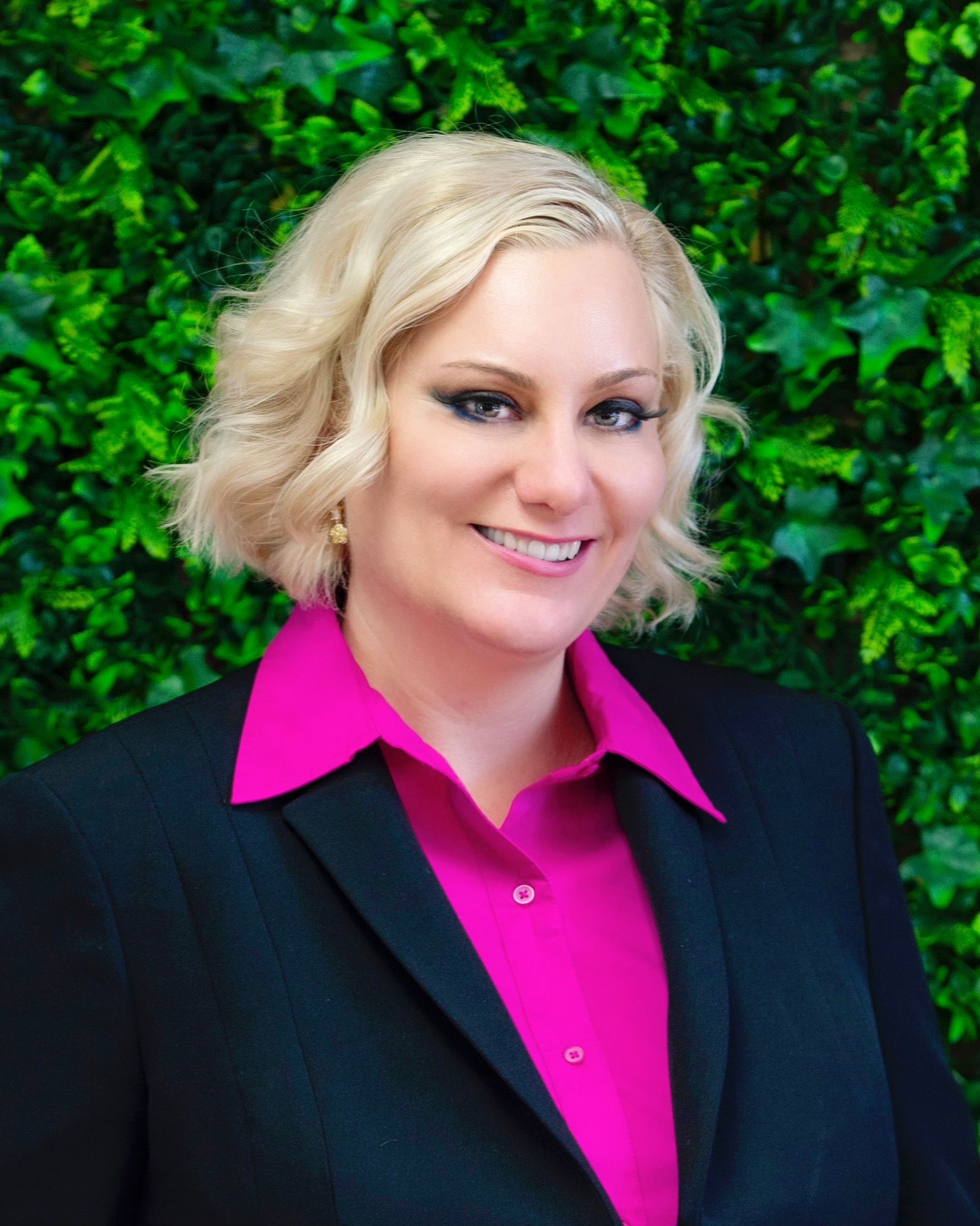 Woman with blonde hair wearing a pink shirt and black blazer smiles in front of green foliage.