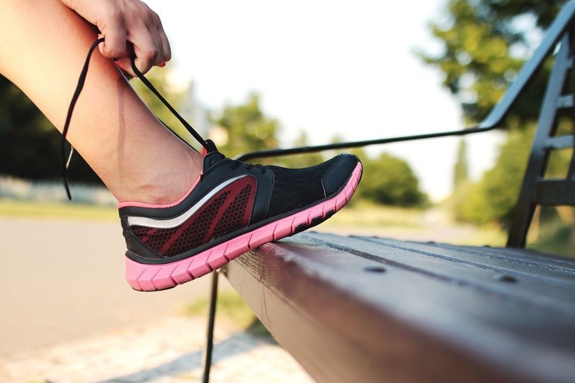 Person tying shoelace on a black and pink running shoe, foot on wooden bench.