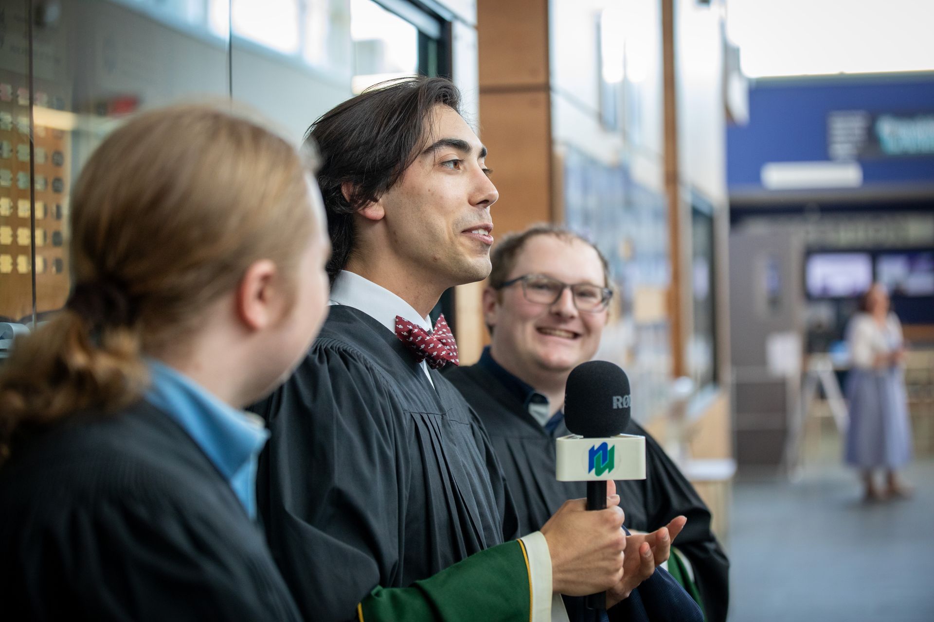 Graduate speaking into a microphone while two people stand beside them outdoors near a building.